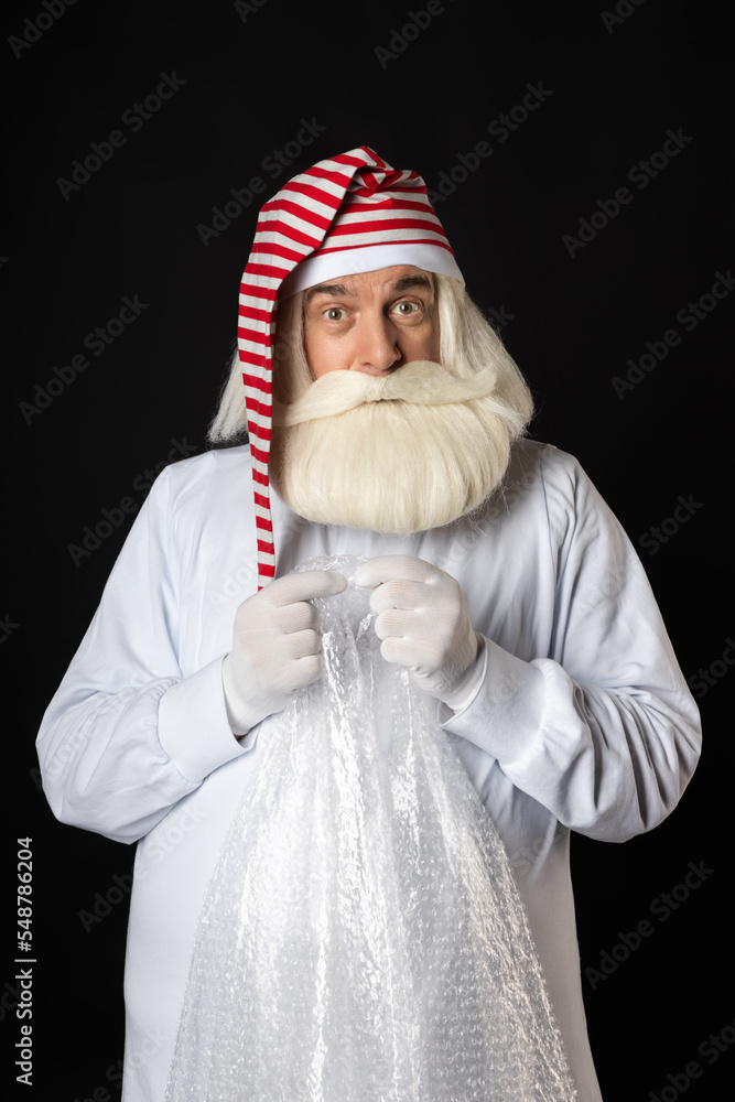 Santa Claus in pajamas and a hat relaxes, presses the bubble wrap in his hands in excitement, squeezing his palms and fingers. The concept of a worried Santa for Christmas, only Black background.