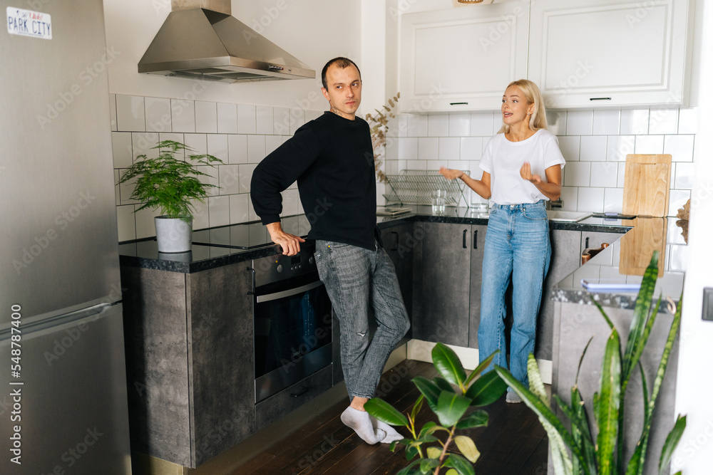Wide shot of angry emotional blonde woman scolding, raising voice, shouting at ignoring tired boyfriend standing in kitchen room looking away. Young couple quarrelling yelling at home. Wide shot of angry emotional blonde woman scolding, raising voice, shouting at ignoring tired boyfriend standing in kitchen room looking away. Young couple quarrelling yelling at home.
