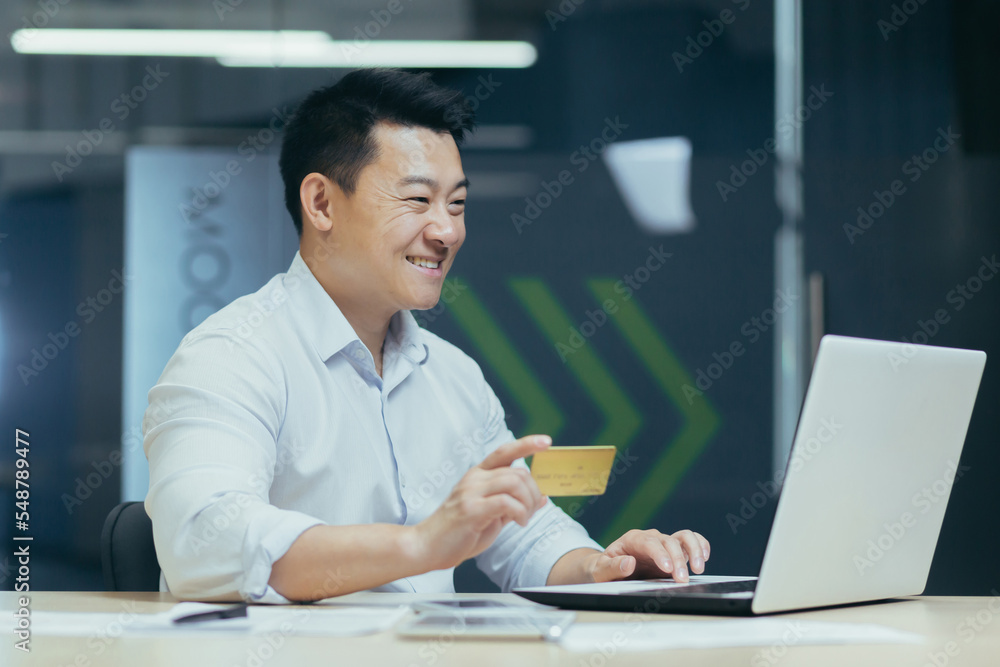 Online shopping. Satisfied young Asian man sitting in the office at the table with a laptop. He holds a credit card in his hands, makes online purchases, orders through the application, smiles. Online shopping. Satisfied young Asian man sitting in the office at the table with a laptop. He holds a credit card in his hands, makes online purchases, orders through the application, smiles.
