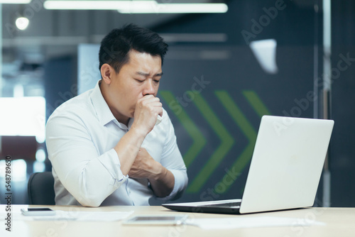 A young Asian freelancer, an office worker fell ill at work. He sits at the table and coughs, covers his mouth with his hand, holds his chest, feels bad. © Liubomir