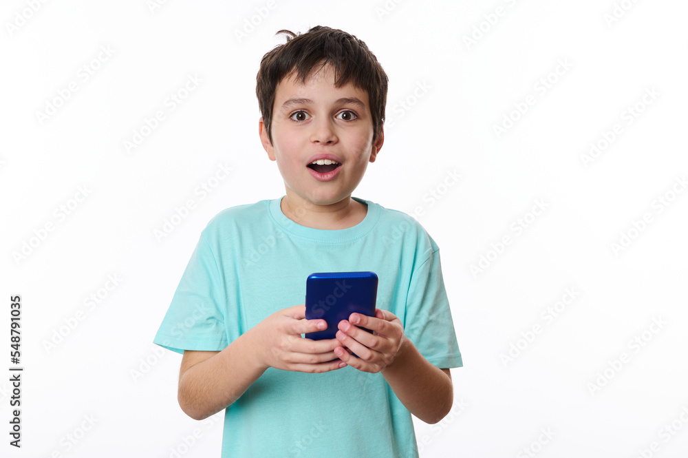Amazed excited cute teenage boy, holding a cell phone and looking at camera with shocked face, child surprised by telephone, online service. Indoor studio shot white background. Wow awesome mobile app Amazed excited cute teenage boy, holding a cell phone and looking at camera with shocked face, child surprised by telephone, online service. Indoor studio shot white background. Wow awesome mobile app