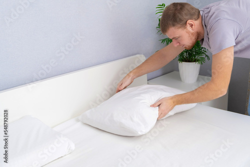 A man tucks a pillow with a fresh bright white pillowcase. Making the bed with fresh bed linen by a white man. The day of the change of bed linen. The day of washing bed linen in the laundry room.