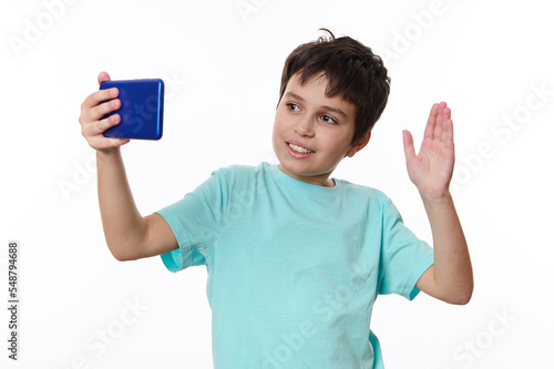 Handsome friendly teenage boy wearing blue t-shirt, using smartphone, smiling and gesturing hi hello to mobile phone. Cute child communicating with parents on video call, isolated on white background