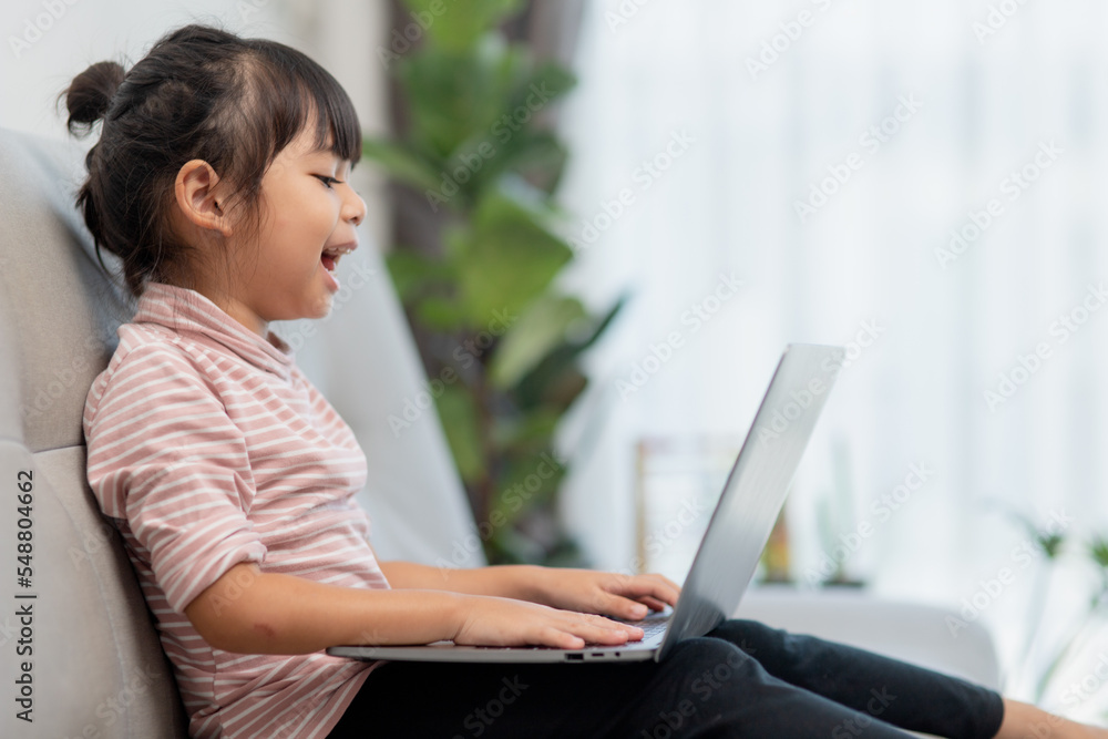 Asian Little school kid girl use laptop computer sitting on sofa alone at home. Child learning reading online social media content, play education lessons game chatting with friends. Asian Little school kid girl use laptop computer sitting on sofa alone at home. Child learning reading online social media content, play education lessons game chatting with friends.