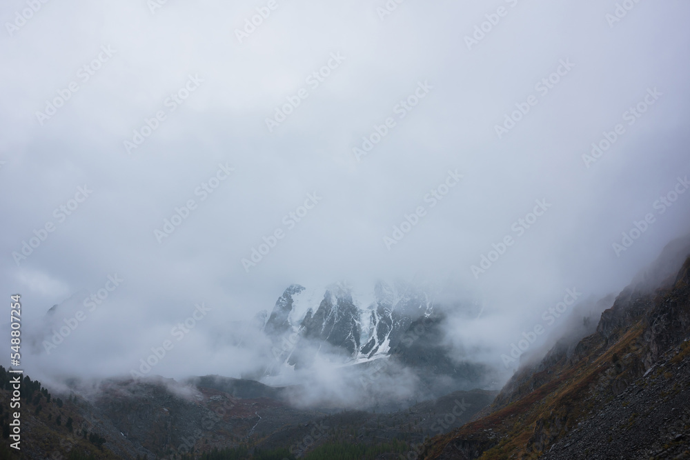 Dark atmospheric landscape with high snow mountains in dense fog in rainy weather. Large snowy mountain range in thick fog in dramatic overcast. Snow-covered rocky mountains in low clouds during rain.