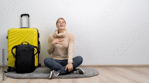 Woman traveler with suitcase in sweater ready to go to travel with tender smile, keeps palm on chest. Passenger travel abroad on weekends getaway.