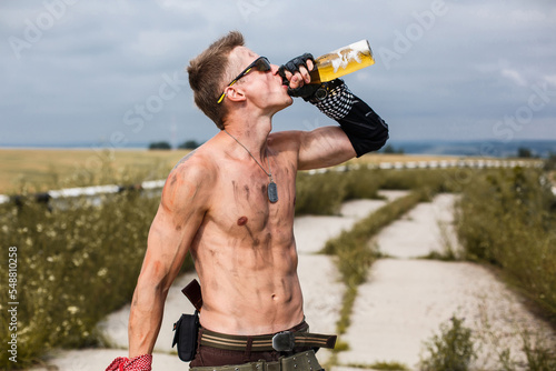 male vandal with dirty naked torso drinks beer, against backdrop of an overgrown abandoned road