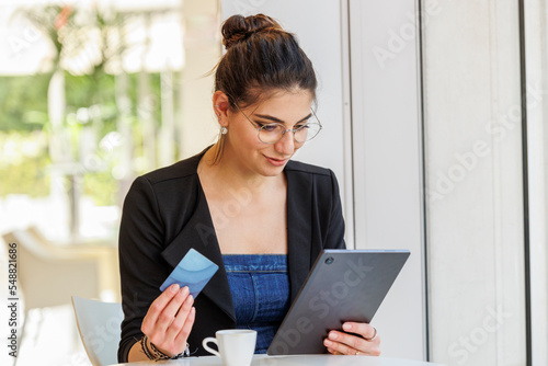 una ragazza mora seduta in un tavolino tiene in mano il proprio tablet  e con una carta di credito è pronta a fare degli acquisti vi internet photo