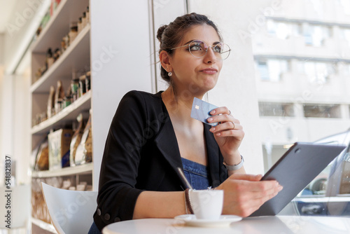 una ragazza mora seduta in un tavolino tiene in mano il proprio tablet  e con una carta di credito è pronta a fare degli acquisti vi internet. L'espressione è di incertezza. photo