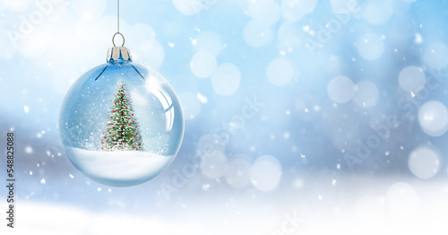 Leinwand Poster Weihnachtskugel mit Weihnachtsbaum hängend vor unscharfem Schneehintergrund
