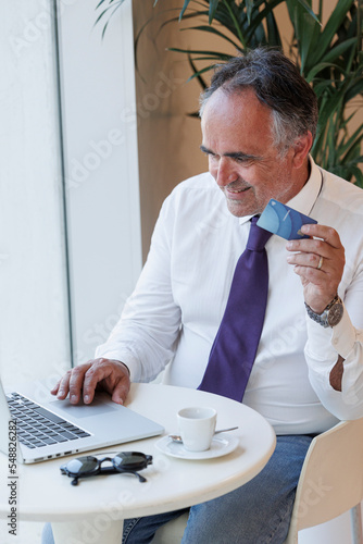 Un signore di mezza età sorridente è seduto al tavolino di un locale mentre guarda il proprio computer portatile e con la carta di credito in mano è pronto a fare degli acquisti via internet . photo