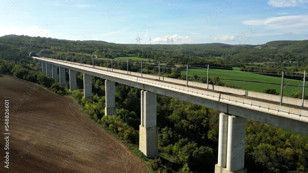 Panoramic view of the railroad tracks for high-speed trains on the high flyover that flows into the tunnel in the mountains. In the background a wind farm generating green energy. Flight from a drone Panoramic view of the railroad tracks for high-speed trains on the high flyover that flows into the tunnel in the mountains. In the background a wind farm generating green energy. Flight from a drone