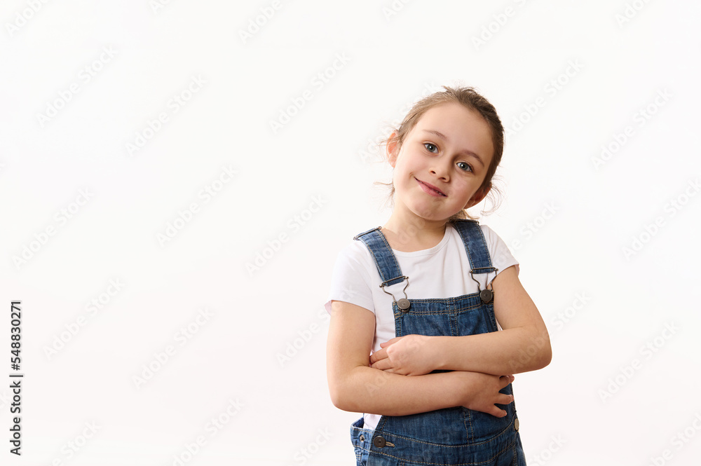 Isolated portrait on white background of adorable little girl, wearing blue denim overalls, hugging herself, cutely smiling looking at camera, isolated over white background. Copy space for promo text Isolated portrait on white background of adorable little girl, wearing blue denim overalls, hugging herself, cutely smiling looking at camera, isolated over white background. Copy space for promo text