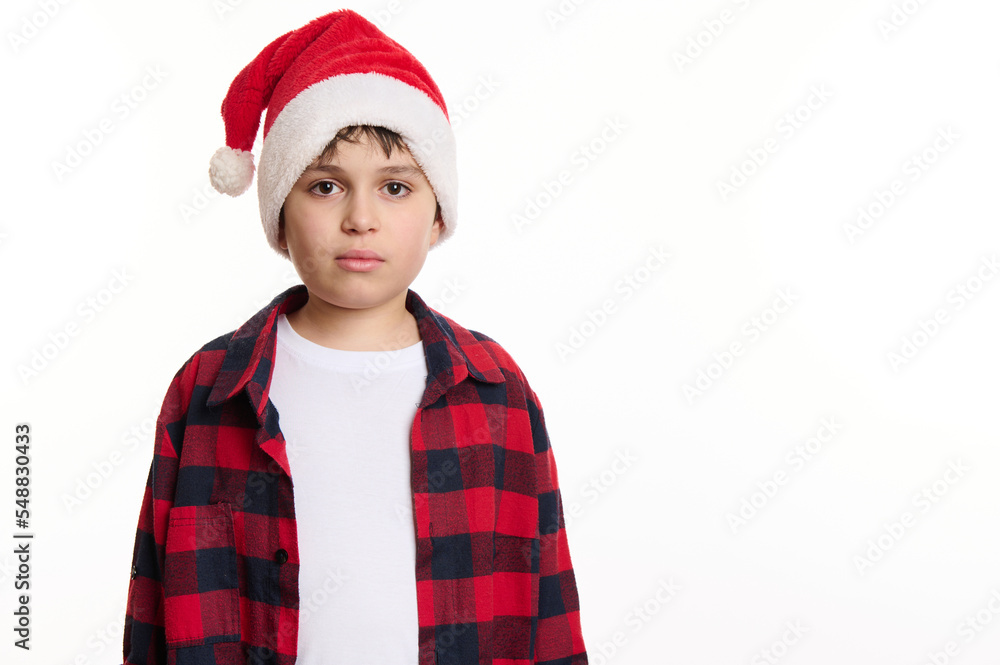 Isolated portrait on white background of a handsome Caucasian preteen boy in Santa hat, in anticipation of Christmas holidays. Pure joy. Winter spirit. New Year party. Copy space for advertising text Isolated portrait on white background of a handsome Caucasian preteen boy in Santa hat, in anticipation of Christmas holidays. Pure joy. Winter spirit. New Year party. Copy space for advertising text