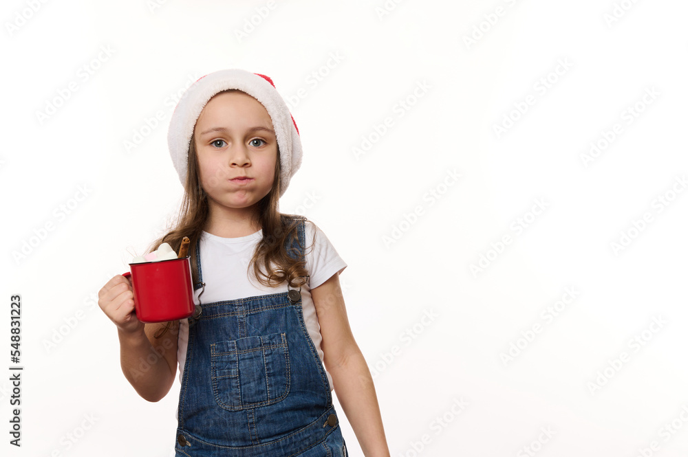 Mischievous 5 years old little girl wearing Santa hat and denim overalls, drinking hot chocolate drink and eating marshmallows, isolated on white background with copy ad space. Christmas atmosphere Mischievous 5 years old little girl wearing Santa hat and denim overalls, drinking hot chocolate drink and eating marshmallows, isolated on white background with copy ad space. Christmas atmosphere