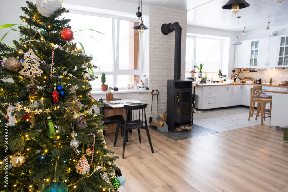 Festive interior of house is decorated for Christmas and New Year in loft style with black stove, fireplace, Christmas tree. Warm studio room with white kitchen, burning wood, cozy and heating of home Festive interior of house is decorated for Christmas and New Year in loft style with black stove, fireplace, Christmas tree. Warm studio room with white kitchen, burning wood, cozy and heating of home