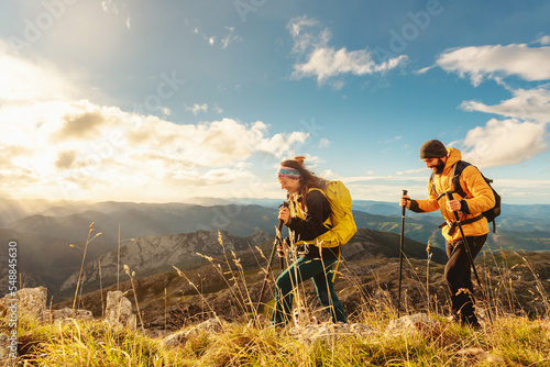 two mountaineers, male and female, trekking up a mountain at sunset. hikers equipped with backpacks, trekking poles and warm clothing walking up the mountain.
