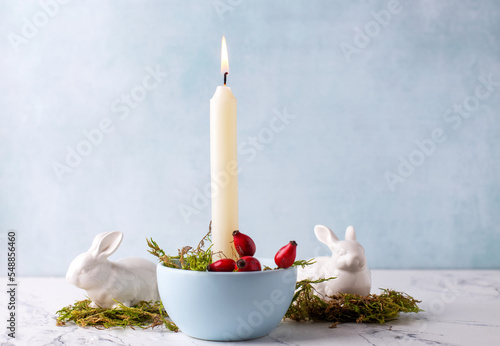 White burning candle, moss, briar berries and white bunnies on white marble background against blue  textured wall. Rabbit is symbool of 2023.. Selective focus. Place for text. photo