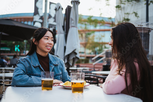 Two laughing young women talking with each other at a bar terrace outdoors. They are having fun © Pablo Rasero