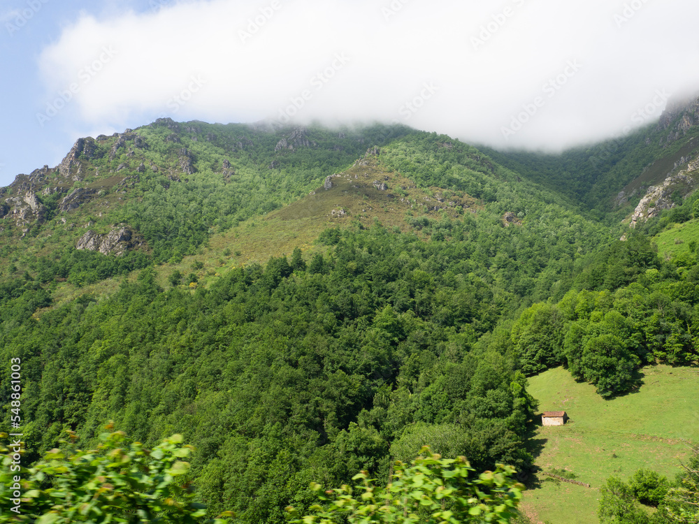 Vistas de una gran extensión de terreno verde con una casita pequeña lejana, en una montaña del norte de España en Asturias, en verano de 2021, al fondo niebla blanca en un día de verano.
