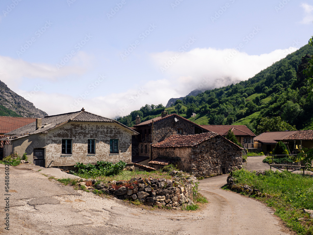 Vistas de las casas del pueblo de  Villar de Vildas, con las montañas verdes al fondo, cielo azul y callejuelas, en verano de 2021 , Asturias, España.