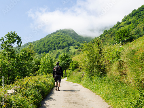 Persona caminando por un sendero solitario, rodeado de naturaleza verde con árboles y montañas al fondo sobre cielo azul y nubes blancas, en verano de 2021 Asturias, España