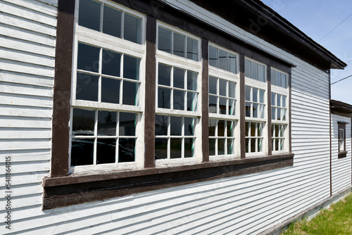 An exterior wooden white vintage building with dark brown trim and a row of windows. The wood wall is made of v-grove clapboard. There are five double hung windows with small square panes of glass. 