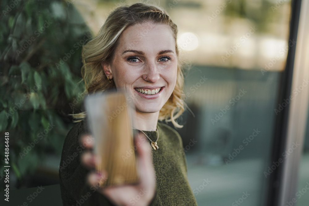 happy smiling young natural alternative looking blond woman in an optimistic & cheerful mood holding or showing mobile phone into the camera pointing to the front with green plants in the background happy smiling young natural alternative looking blond woman in an optimistic & cheerful mood holding or showing mobile phone into the camera pointing to the front with green plants in the background