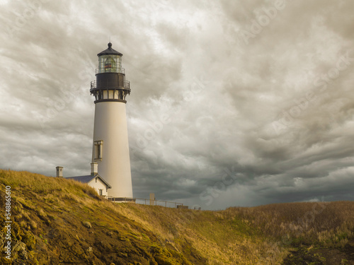 A white high lighthouse on the green shore of the ocean against the background of a gloomy sky with thunderclouds. Marine navigation, beautiful structure, architecture, landscape, tourism.