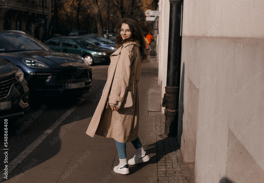 Young woman in a coat outside. Outdoor autumn portrait of young elegant fashionable woman wearing trendy glasses, camel color coat, shoulder bag, walking in street of European city. Copy space