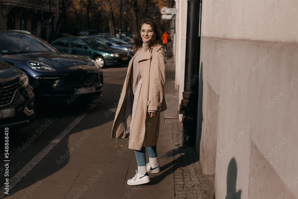 Fototapeta premium Young woman in a coat outside. Outdoor autumn portrait of young elegant fashionable woman wearing trendy glasses, camel color coat, shoulder bag, walking in street of European city. Copy space