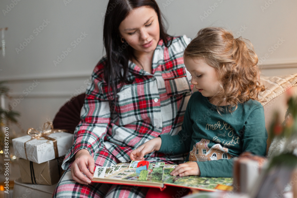 Fototapeta premium Mom and daughter sit on the couch with toys and read a book on Christmas Eve. Happy family holidays
