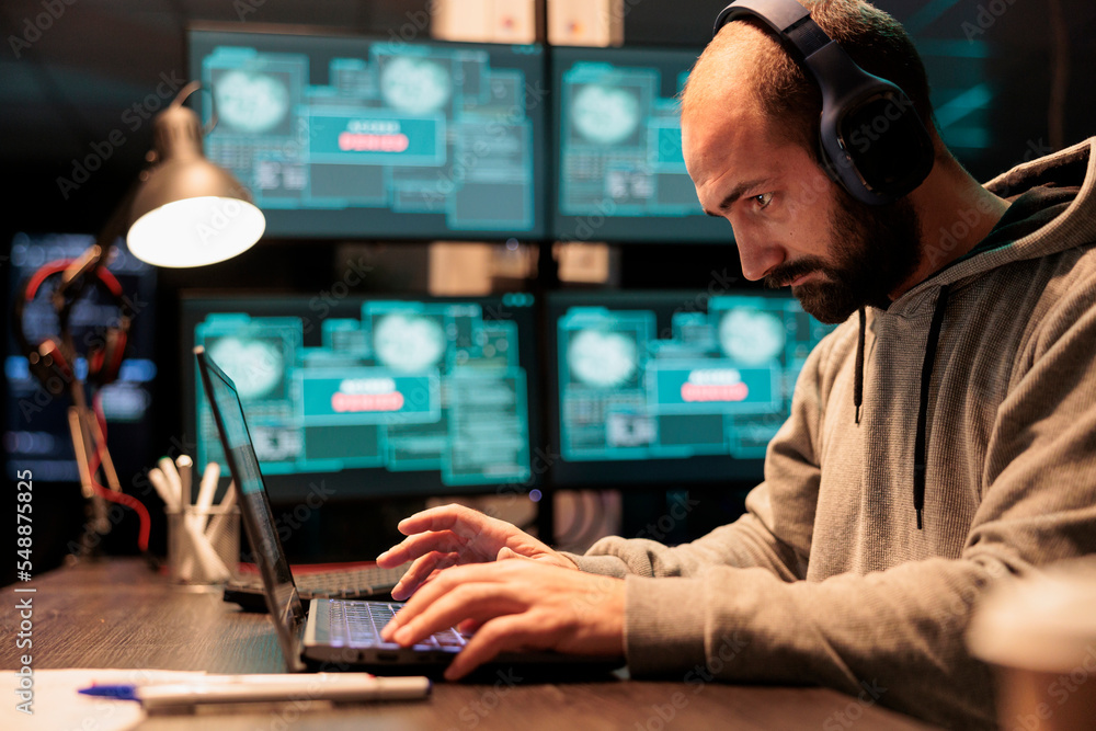 Cyber criminal hacking security system to steal information, wearing headphones at office. Coding virus to create malware and break into database servers, using multiple monitors. Cyber criminal hacking security system to steal information, wearing headphones at office. Coding virus to create malware and break into database servers, using multiple monitors.