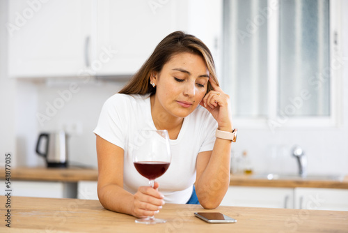 Young woman with glass of red wine in kitchen looks sadly at screen of a mobile phone