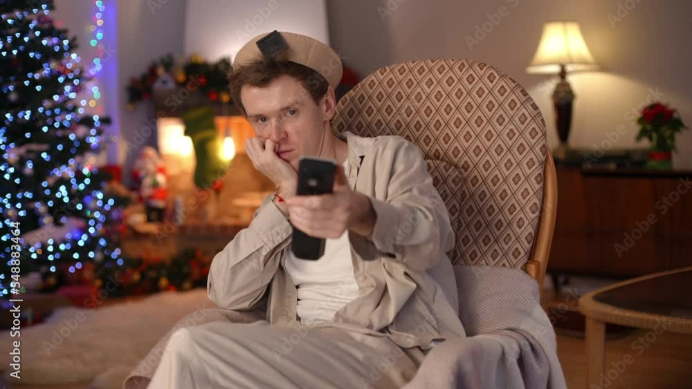Portrait of sad bored LGBT man looking at camera talking turning off TV with remote control sighing looking away. Lonely sad Caucasian guy at home indoors on Christmas eve. New Year and lifestyle Portrait of sad bored LGBT man looking at camera talking turning off TV with remote control sighing looking away. Lonely sad Caucasian guy at home indoors on Christmas eve. New Year and lifestyle
