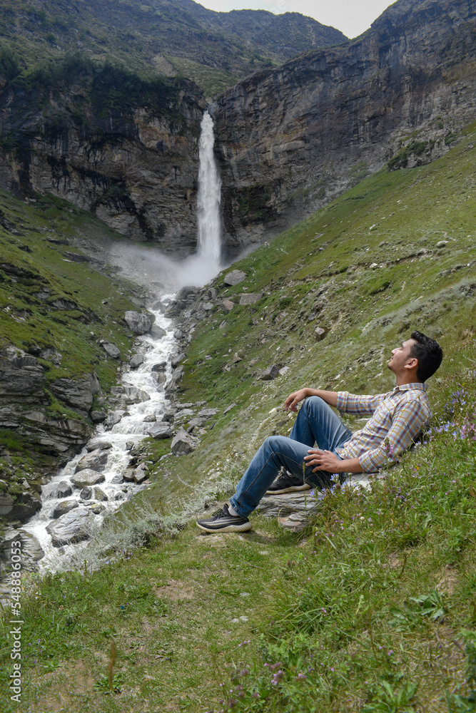 Guy enjoying sitting on grass hill slope in daylight with beautiful tall waterfall in background
