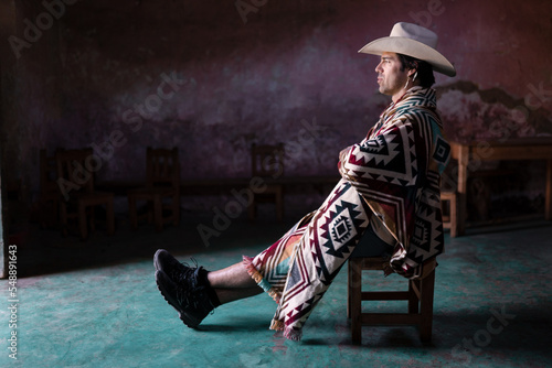 A young Mexican is sitting with crossed arms inside of a church wearing a hat and a poncho