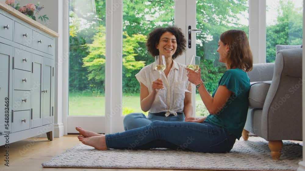 Same sex female couple or friends at home sitting on floor in lounge drinking wine and making a toast together - shot in slow motion Same sex female couple or friends at home sitting on floor in lounge drinking wine and making a toast together - shot in slow motion