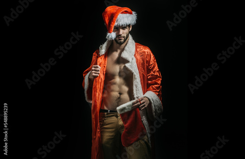 Christmas sexy man. Strong guy in santa hat. New year strip and gifts for adults. Muscle gay at xmas. Santa with muscular body. Sexy young Santa Claus on black background. photo