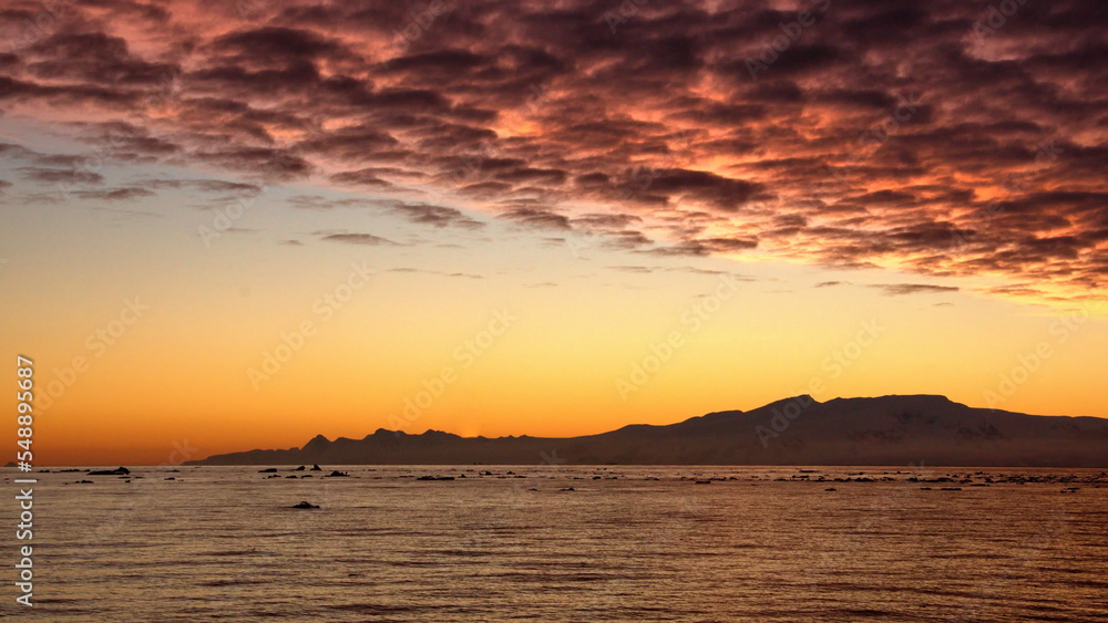 Pink clouds in an orange sky, with mountains and icebergs in silhouette, at Cierva Cove, Antarctica, at sunset