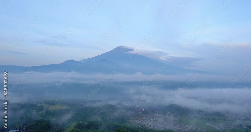 Aerial shot over Indonesia countryside shrouded in sea of mist with huge mountain on the background in the morning - Sumbing Mountain, Central Java, Indonesia Aerial shot over Indonesia countryside shrouded in sea of mist with huge mountain on the background in the morning - Sumbing Mountain, Central Java, Indonesia