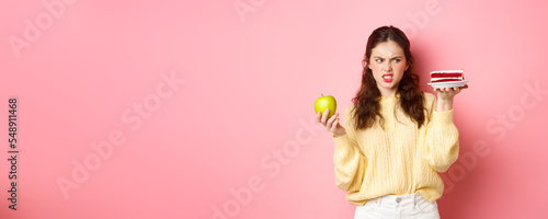 Healthy girl on diet looks with disgust at piece of cake, holding green apply, grimacing dissatisfied, standing against pink background