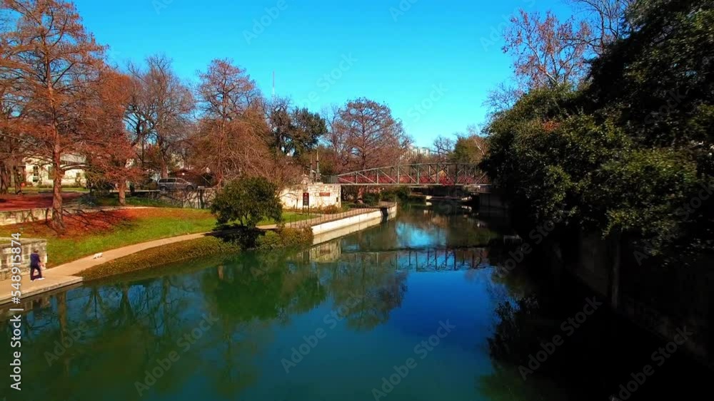 Aerial: Johnson Street Pedestrian Bridge Amidst Bare Trees In Park, Drone Flying Backwards Over River On Sunny Day - San Antonio, Texas Aerial: Johnson Street Pedestrian Bridge Amidst Bare Trees In Park, Drone Flying Backwards Over River On Sunny Day - San Antonio, Texas