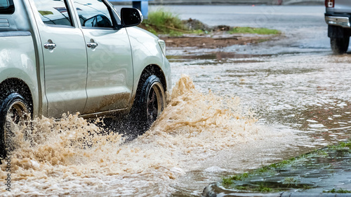 Pickup truck passing through flooded road. Driving car on flooded road during flood caused by torrential rains. Flooded city road with large puddle. Splash by car through flood water. Selective focus.