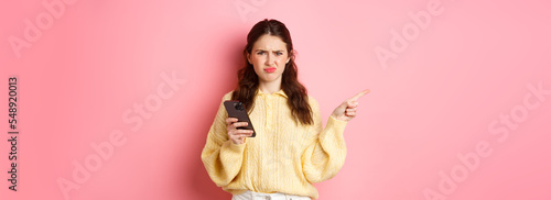 Displeased girl grimacing, frowning upset and pointing finger aside at left copyspace while holding smartphone, standing over pink background