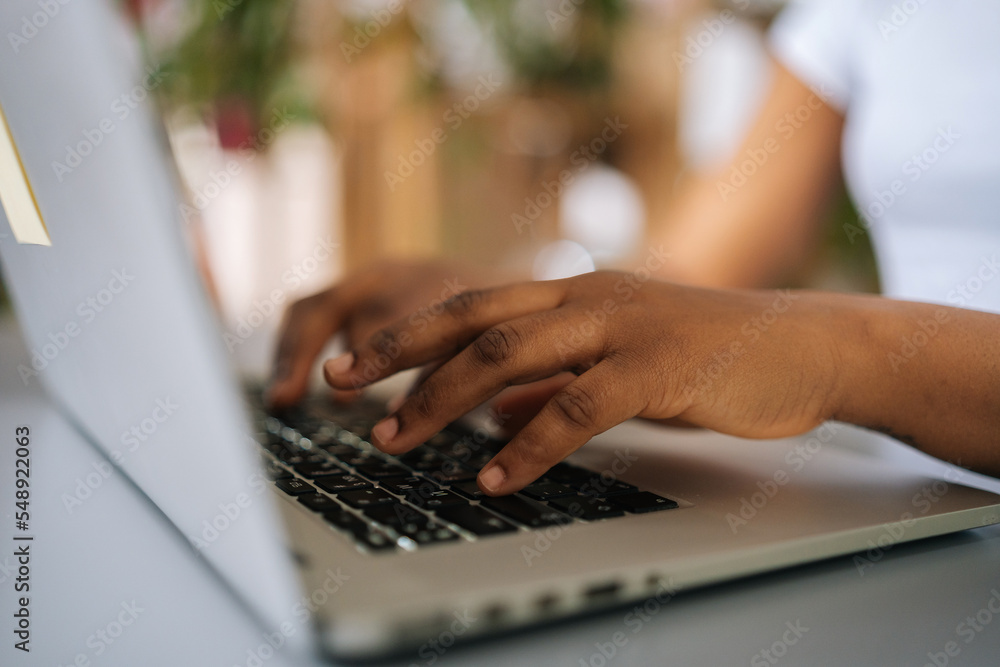Close-up side view of unrecognizable African-American business woman typing on laptop keyboard sitting at home office desk, on beautiful blurry background. Close-up side view of unrecognizable African-American business woman typing on laptop keyboard sitting at home office desk, on beautiful blurry background.