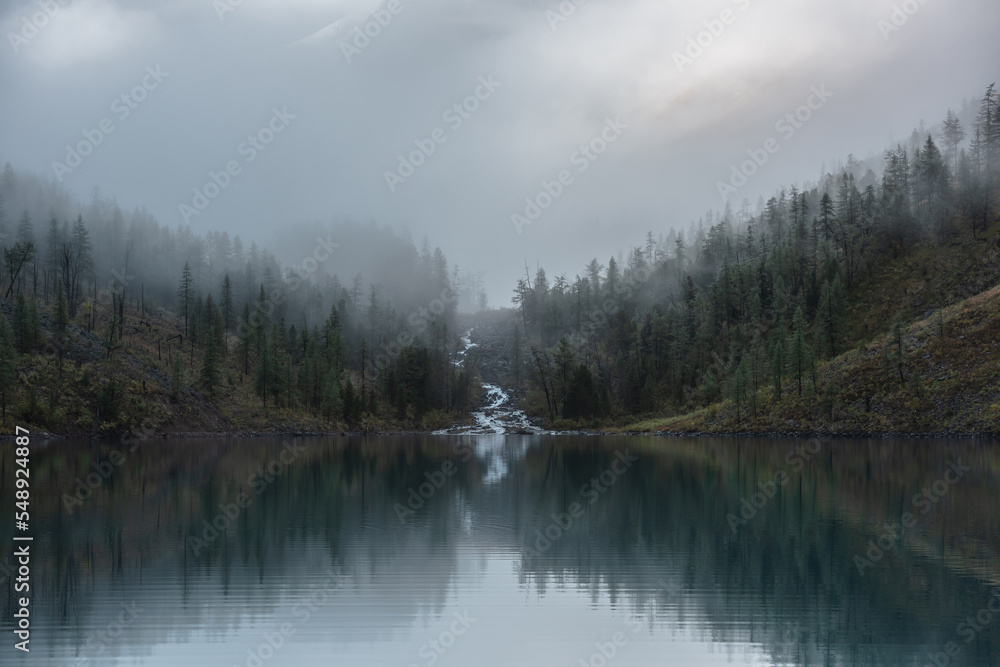 Obraz premium Mountain creek flows from forest hills into glacial lake in mysterious fog. Small river and coniferous trees reflected in calm alpine lake in early morning. Tranquil misty scenery with mountain lake.