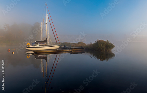 Foggy morning in small river port with anchored sailboats at wooden pier, due...