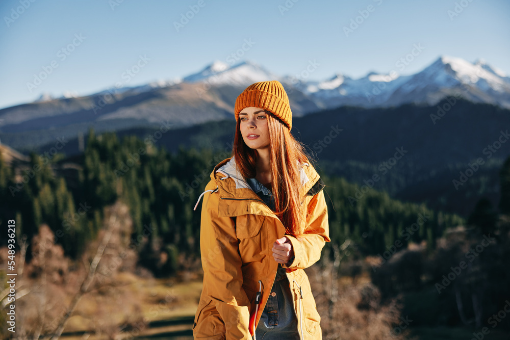 Woman hiking in the mountains in the fall with a smile with teeth and happiness in a yellow cape with red hair full-length stands against the backdrop of trees and mountains in the sunset light Woman hiking in the mountains in the fall with a smile with teeth and happiness in a yellow cape with red hair full-length stands against the backdrop of trees and mountains in the sunset light