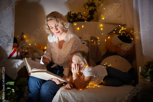 Cute mother and daughter reeading book in children room with Christmas garlands. Tradition of decorating house and dressing up for the holidays. Gifts for the family. Happy childhood and motherhood photo